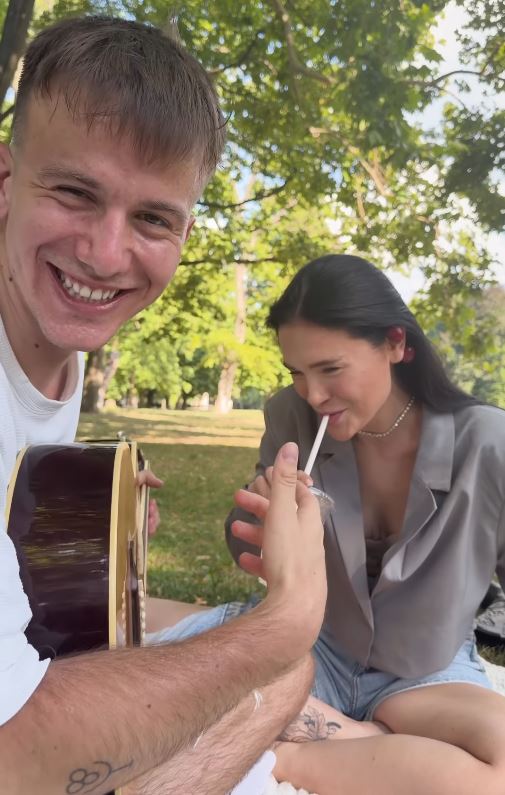 Dvojica relaxuje v parku, muž hrá na gitare, žena sa usmieva a pije cez slamku.