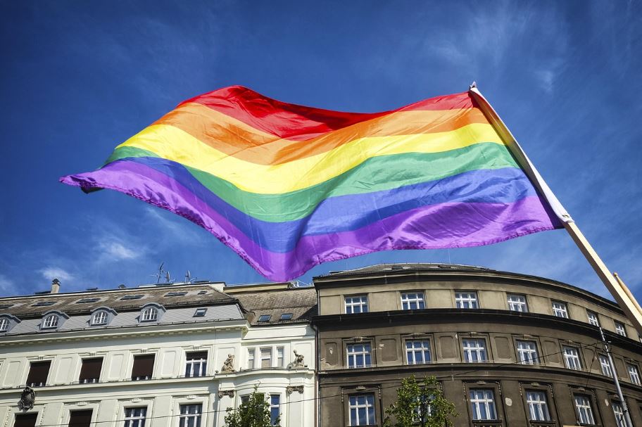 Dúhová vlajka vejúca nad mestskými budovami symbolizuje podporu LGBTQ+ komunity pod modrou oblohou.