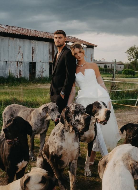 Pár v elegantnom oblečení pózuje s viacerými veľkými psami pred starou kovovou budovou na vidieku.
