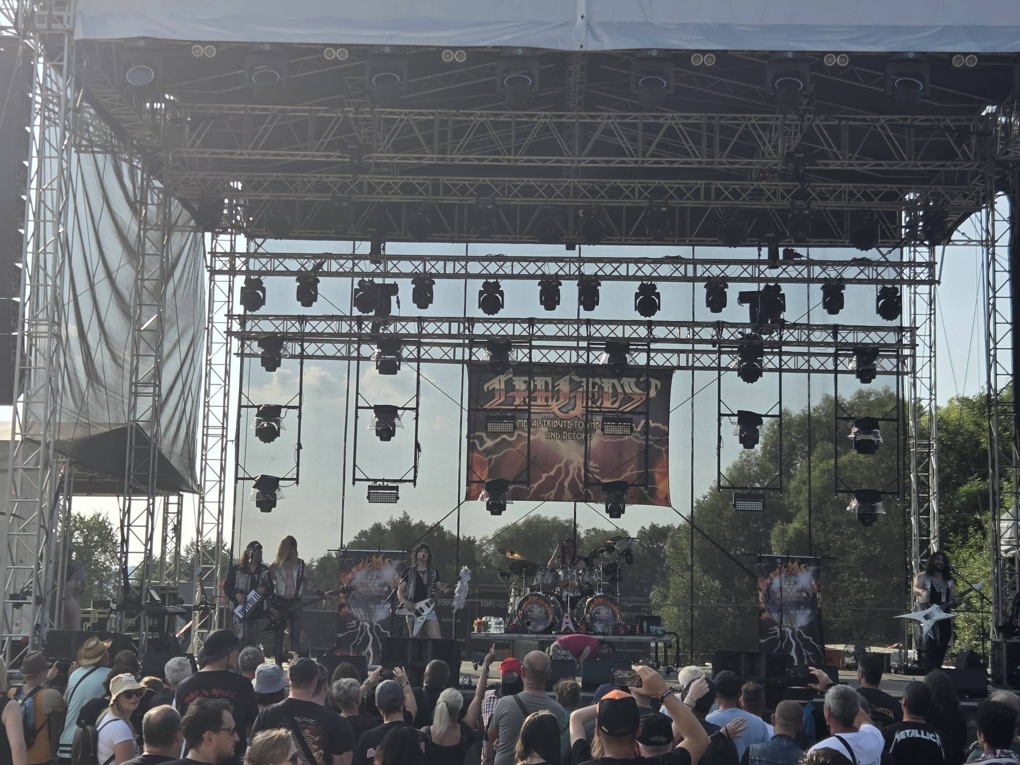 Koncert rockovej kapely na otvorenom pódiu s veľkým publikom, nasvietenie a záves s logom kapely v pozadí.