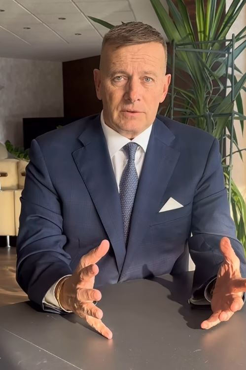 Oblečený muž v obleku pri stole gestikuluje, za ním rastlina a pohovka v modernej kancelárii.