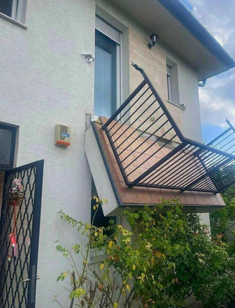 Spadnutý balkón na rodinnom dome s kovovým zábradlím, poškodená strecha a okolie s rastlinami.
