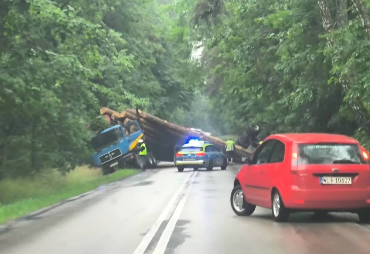 Nehoda nákladného auta s drevom blokuje cestu v lese, policajné auto na mieste, červené auto čaká.