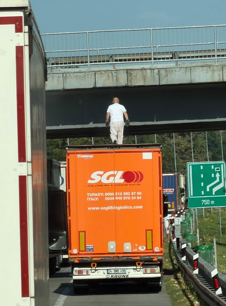 Muž stojí na streche oranžového návesu kamióna pod mostom na diaľnici.