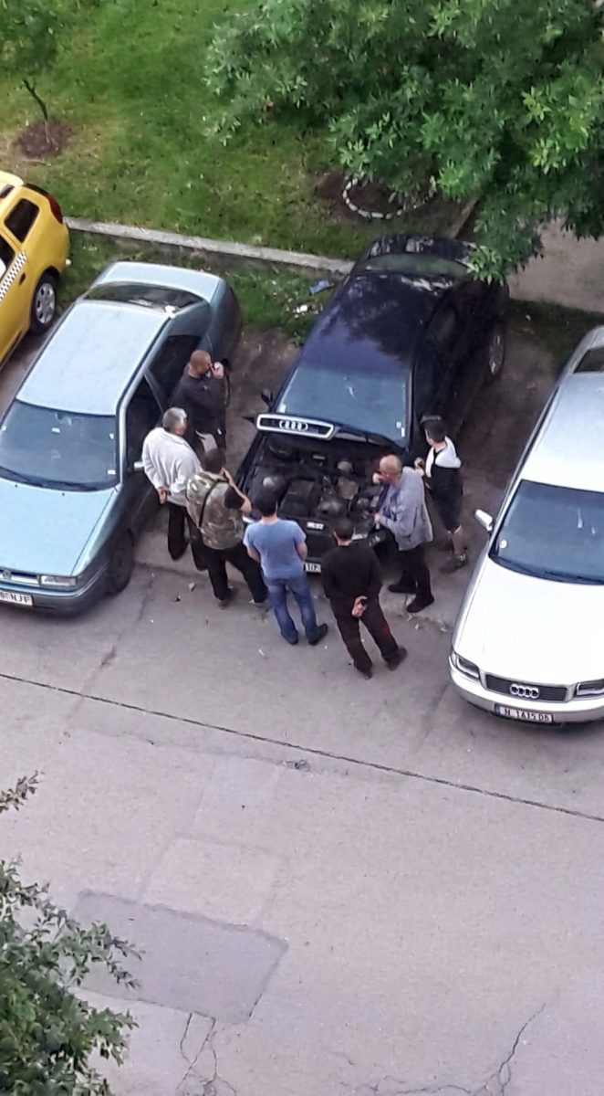 Mužská skupina diskutujúca nad otvorenou kapotou auta na parkovisku medzi zaparkovanými vozidlami.