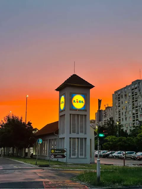 Večerný výhľad na Lidl vežu pred oranžovou oblohou v mestskom prostredí s parkovanými autami.