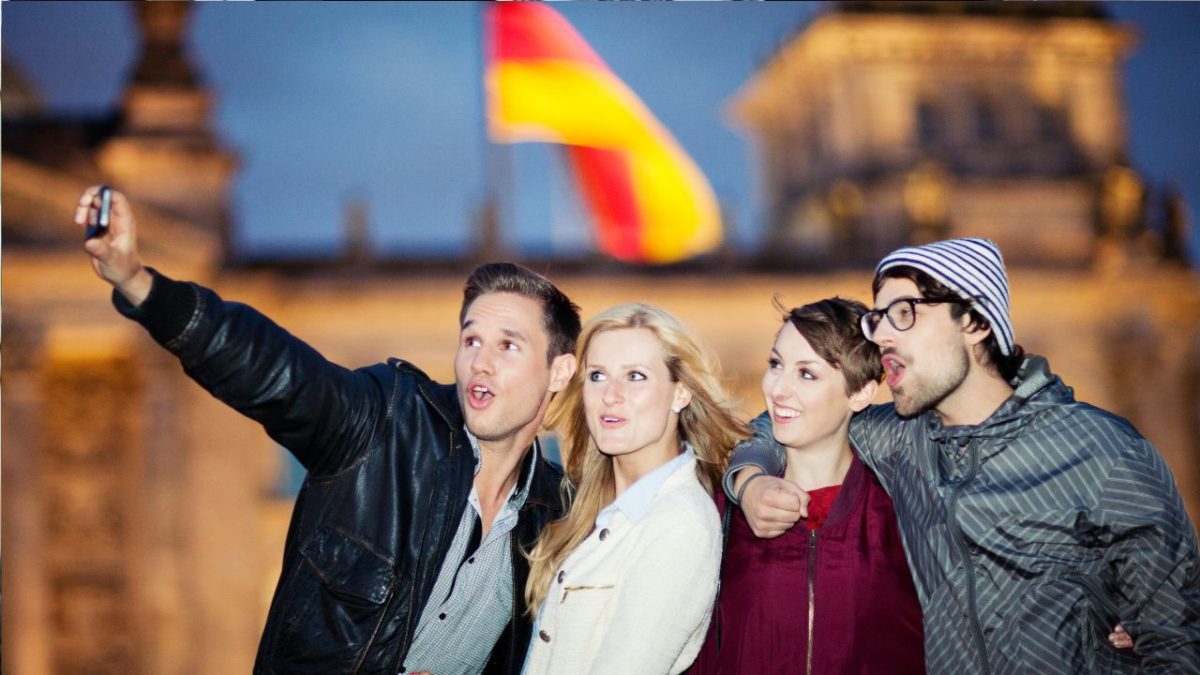 Skupina mladých ľudí si robí selfie pred nemeckou vlajkou, Berlín na pozadí.