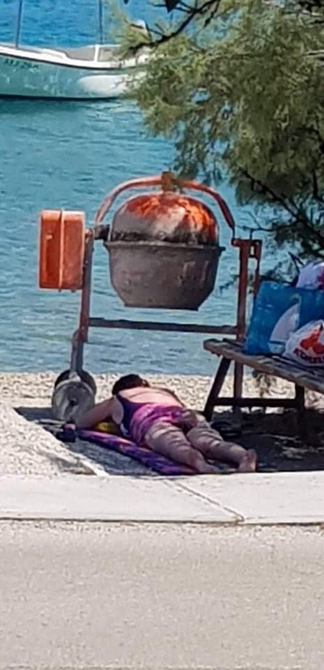 Osoba ležiaca na brehu pri stavebných nástrojoch, v pozadí je modrá voda a loď.