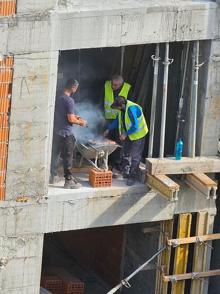 Robotníci na stavbe pripravujú obed na provizórnom grile z fúrika, obklopení stavebným materiálom.