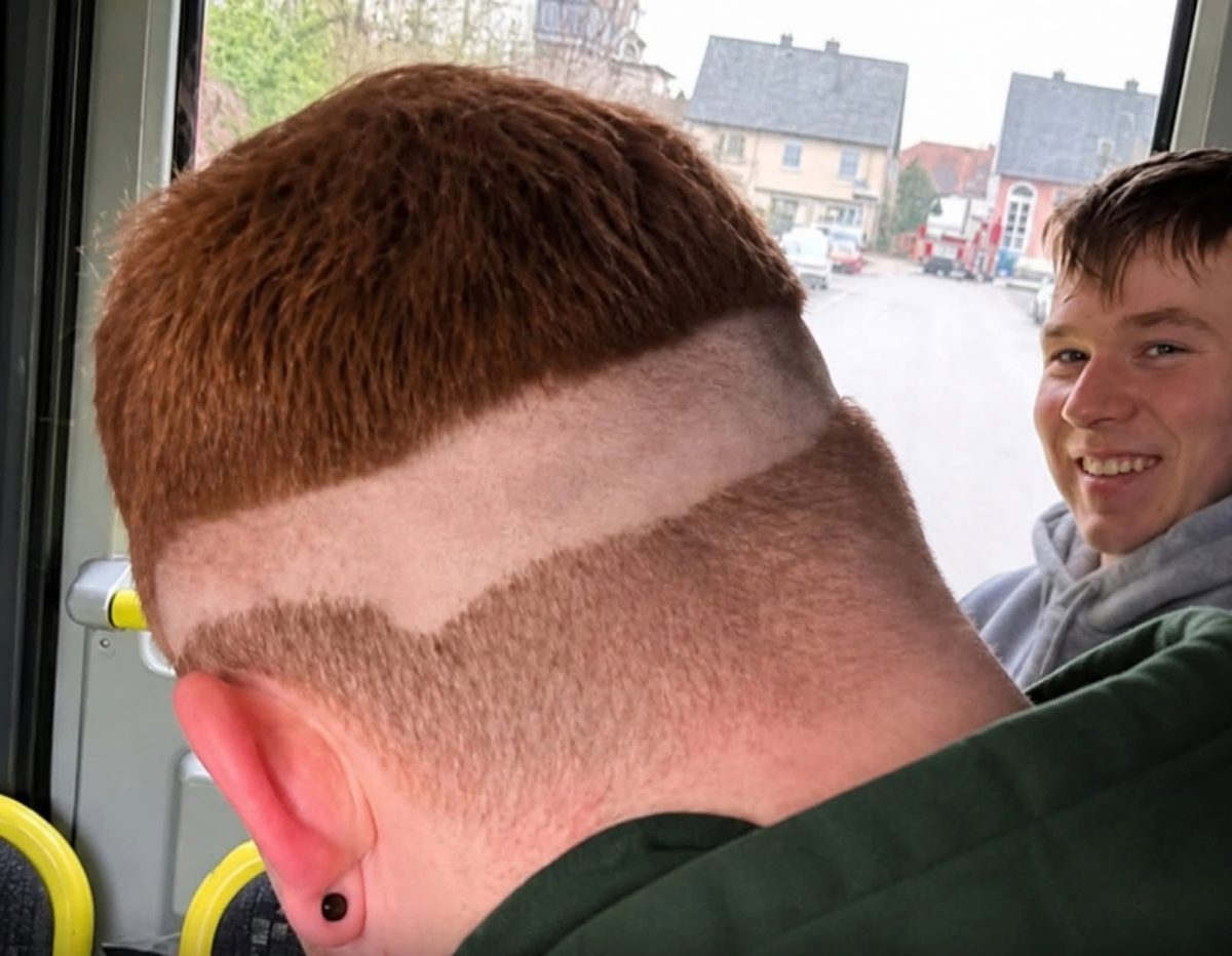 Osoba s neobvyklým účesom v autobuse, veľký úsmev ďalšej osoby v pozadí.
