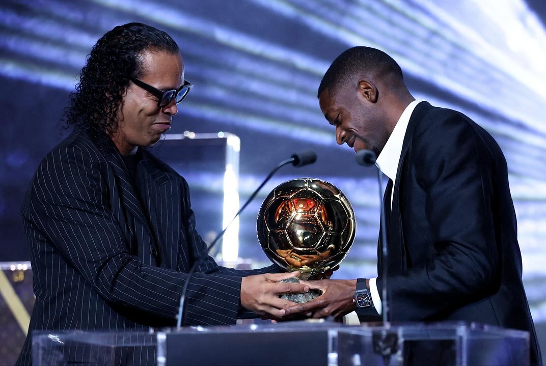 Dvaja muži v oblekách na pódiu si navzájom odovzdávajú zlatú futbalovú trofej.