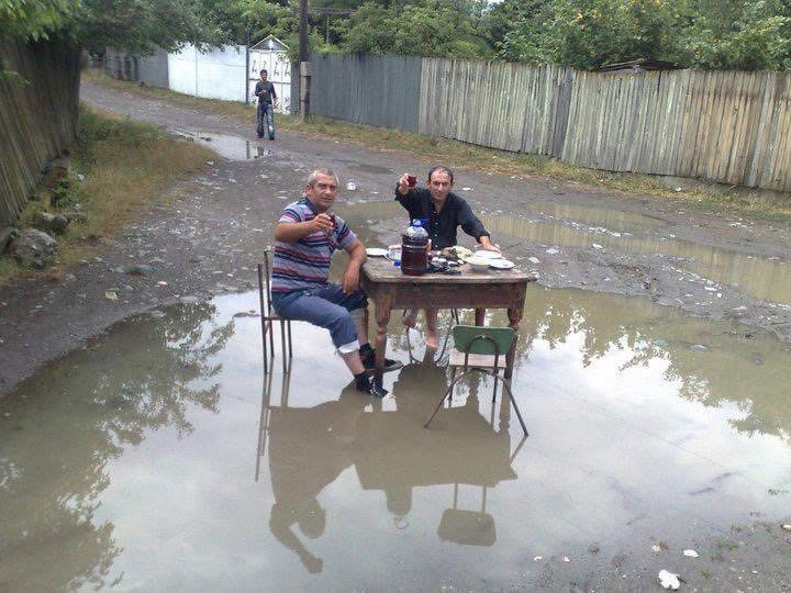 Dve osoby sedia pri stole uprostred kaluže na nespevnenej ceste a pijú nápoje, zatiaľ čo ich obklopuje jedlo.