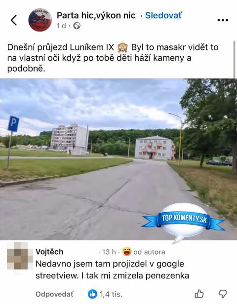 Cesta v obytnom sídlisku s panelákmi, ľavostranné parkovisko, trávnik a stromy na pravej strane.
