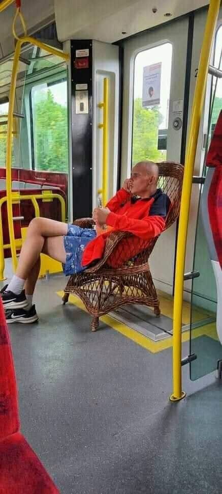 Muž sedí v ratanovom kresle v električke, oblečený v červenom saku a modrých kraťasoch, hovorí do mobilu.
