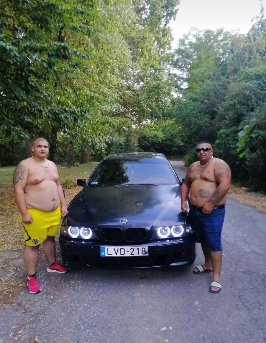 Dvaja muži pri čiernom BMW zaparkovanom na ceste, obklopení zeleňou.