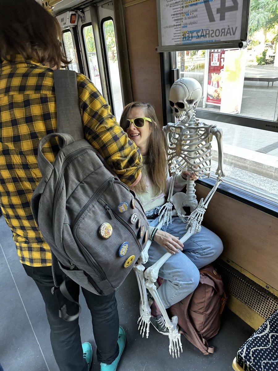 Osoba v autobuse drží kostru na kolenách, usmieva sa a nosí slnečné okuliare. Druhá postava s batohom stojí.