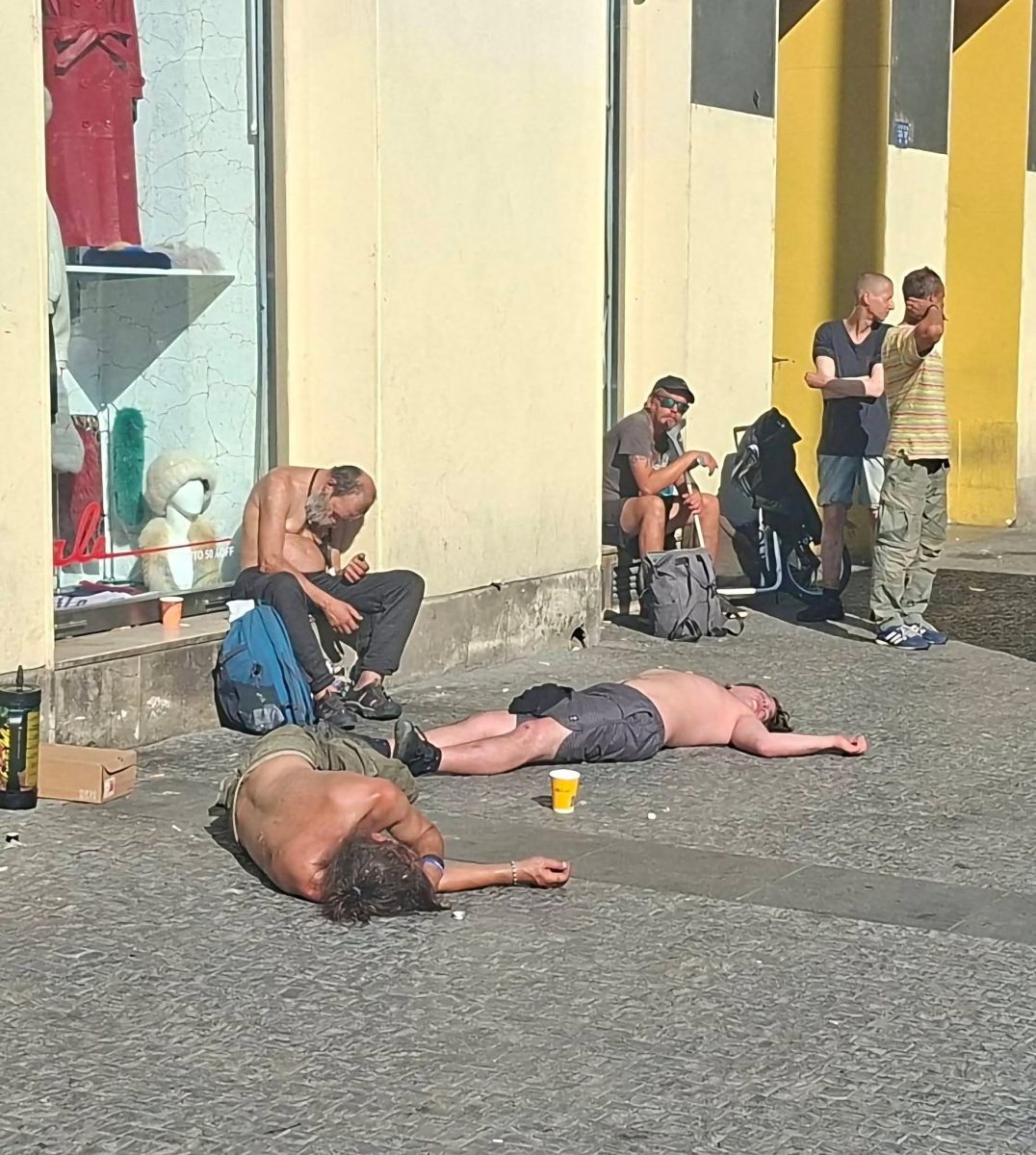 Ľudia odpočívajúci alebo čakajúci v tieni budovy na slnečnej ulici.