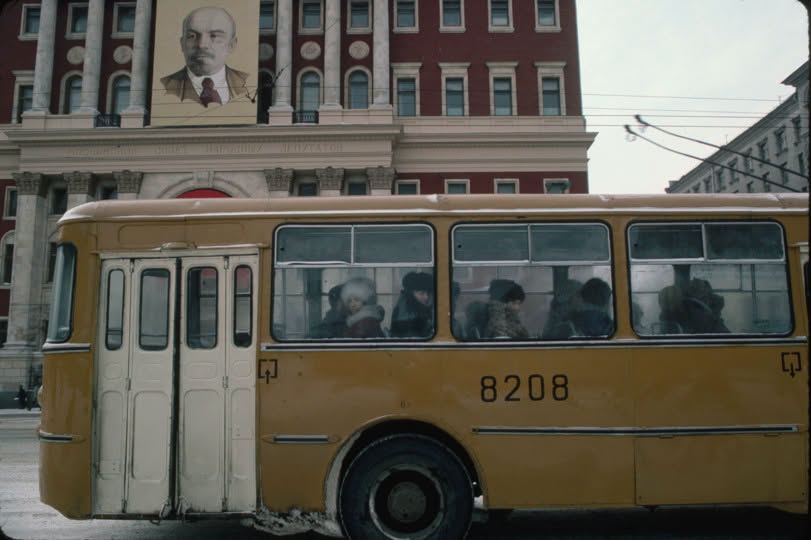 Žltý autobus plný cestujúcich prechádza okolo historickej budovy s veľkým portrétom na hlavnej ulici mesta.