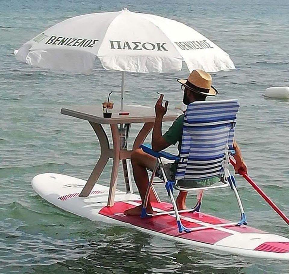 Muž relaxuje na paddleboarde s ležadlom a dáždnikom, plávajúci na mori s drinkom. Letná pohoda a originalita na vode.