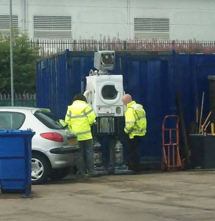 Dvaja muži v reflexných bundách skúmajú práčku umiestnenú na robotickom podstavci pri modrom kontajneri.