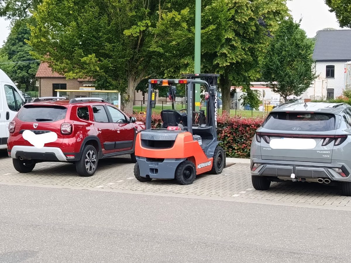 Červené a sivé autá parkujúce vedľa vysokozdvižného vozíka na parkovisku pri ceste.