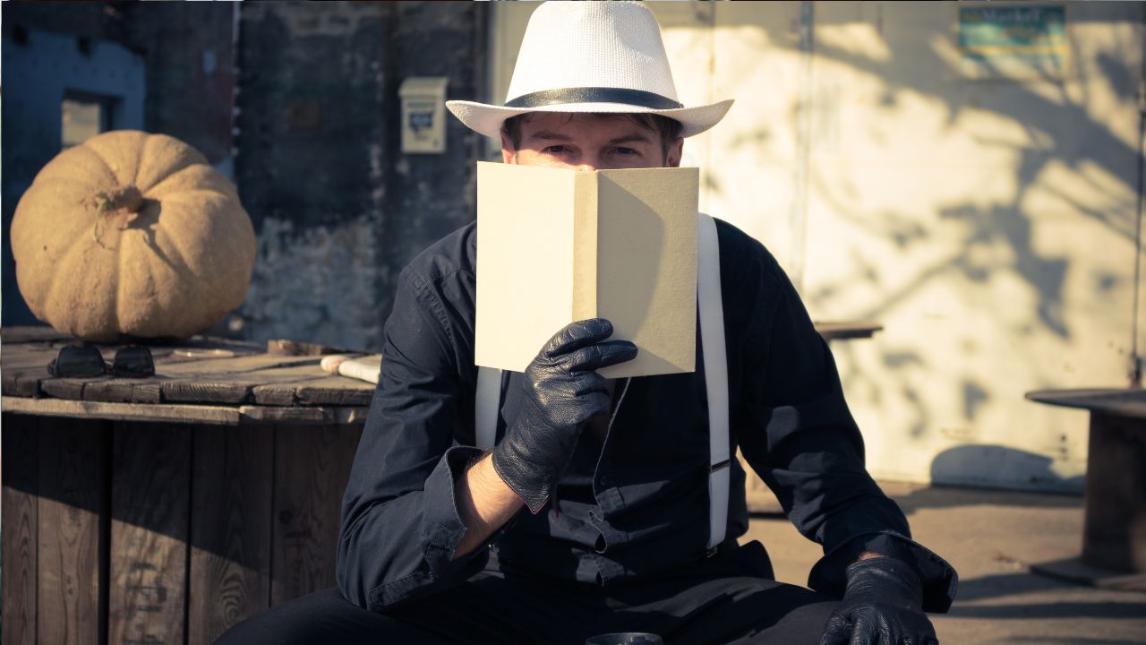 Muž v klobúku drží knihu pred tvárou, sedí vonku pri stole s veľkou tekvicou.