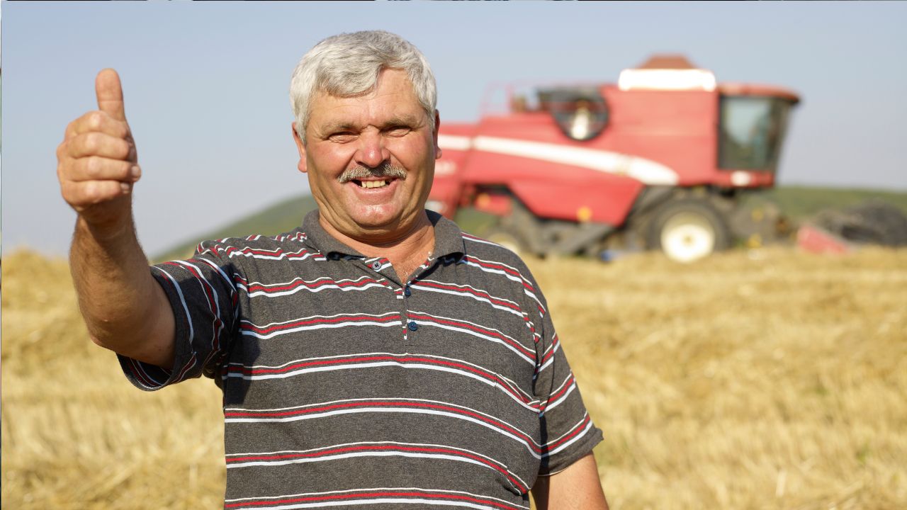 Farmár v poli s kombajnom v pozadí, usmievajúci sa s palcom hore, symbolizuje úspešnú žatvu.