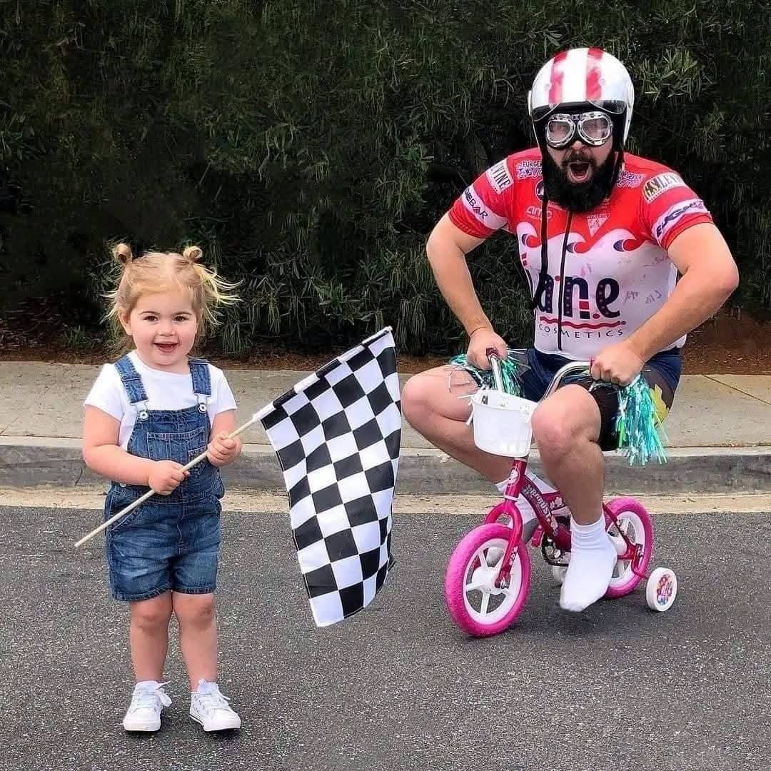 Muž na malom bicykli s detskou prilbou, dieťa s kockovanou vlajkou na štarte. Humor a radosť v parku.