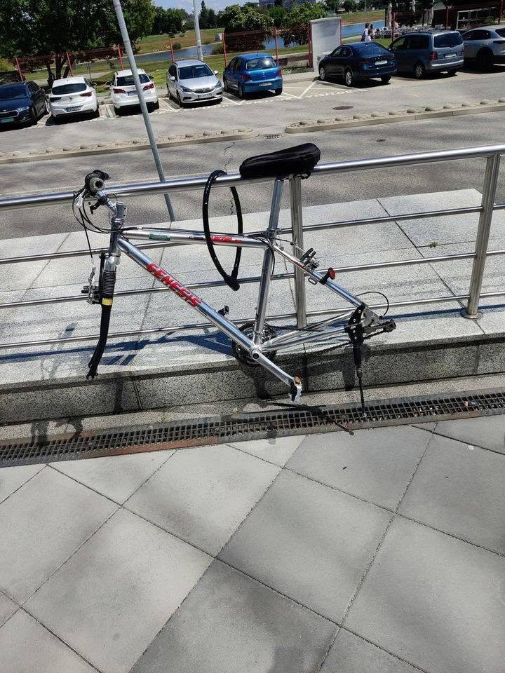 Neúplný bicykel pripevnený k zábradliu pri parkovisku, chýbajúce kolesá.