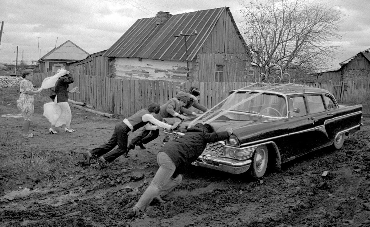 Skupina ľudí tlačí auto zapadnuté v blate pred dreveným domom, žena v svadobných šatách kráča v blízkosti.