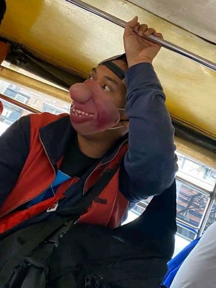Osoba v autobuse s vtipnou maskou, ktorá zobrazuje smiešnu tvár. Drží sa madla nad hlavou.