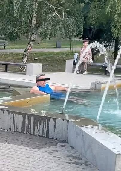 Muž relaxuje v mestskom fontáne počas teplého dňa; ľudia sedia na laviciach v pozadí.