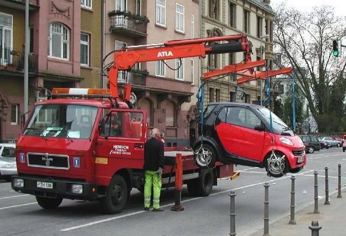 Červený odťahový voz preváža malé auto na rušnej ulici pred historickými budovami.
