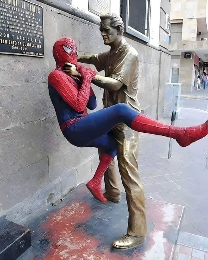 Socha objíma muža v kostýme pavúčieho superhrdinu na ulici, netradičný moment v meste.