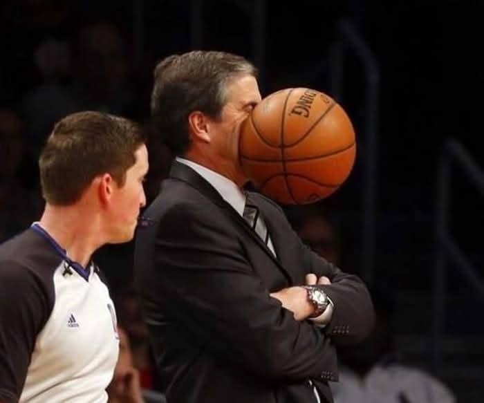 Basketbalová lopta vráža do muža v obleku počas zápasu, s rozhodcom stojacim neďaleko.