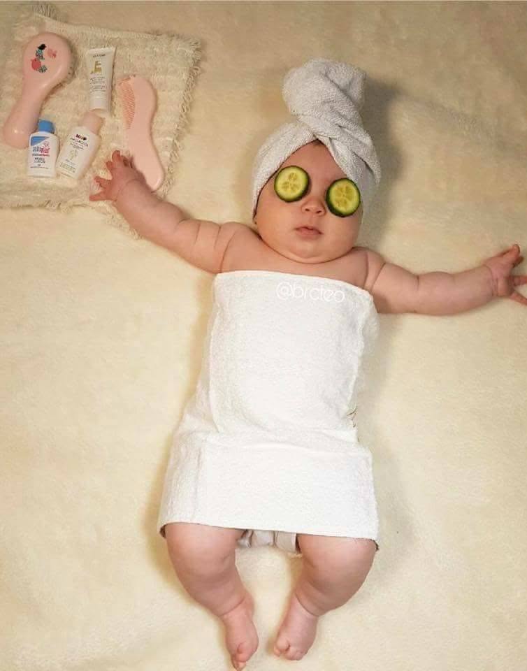 Dieťa v bielom uteráku a so zaviazanou hlavou si užíva relax s plátkami uhorky na očiach.
