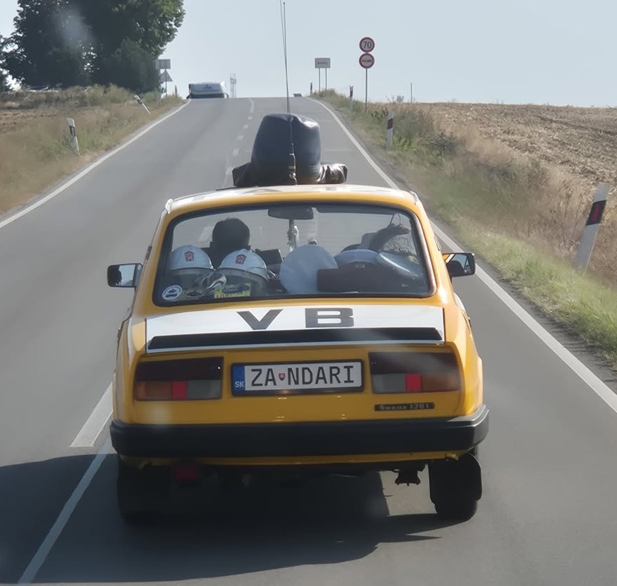 Žlté auto Škoda na vidieckej ceste, na streche maják a nápis VB, presúvajúce sa po mierne vyvýšenom teréne.
