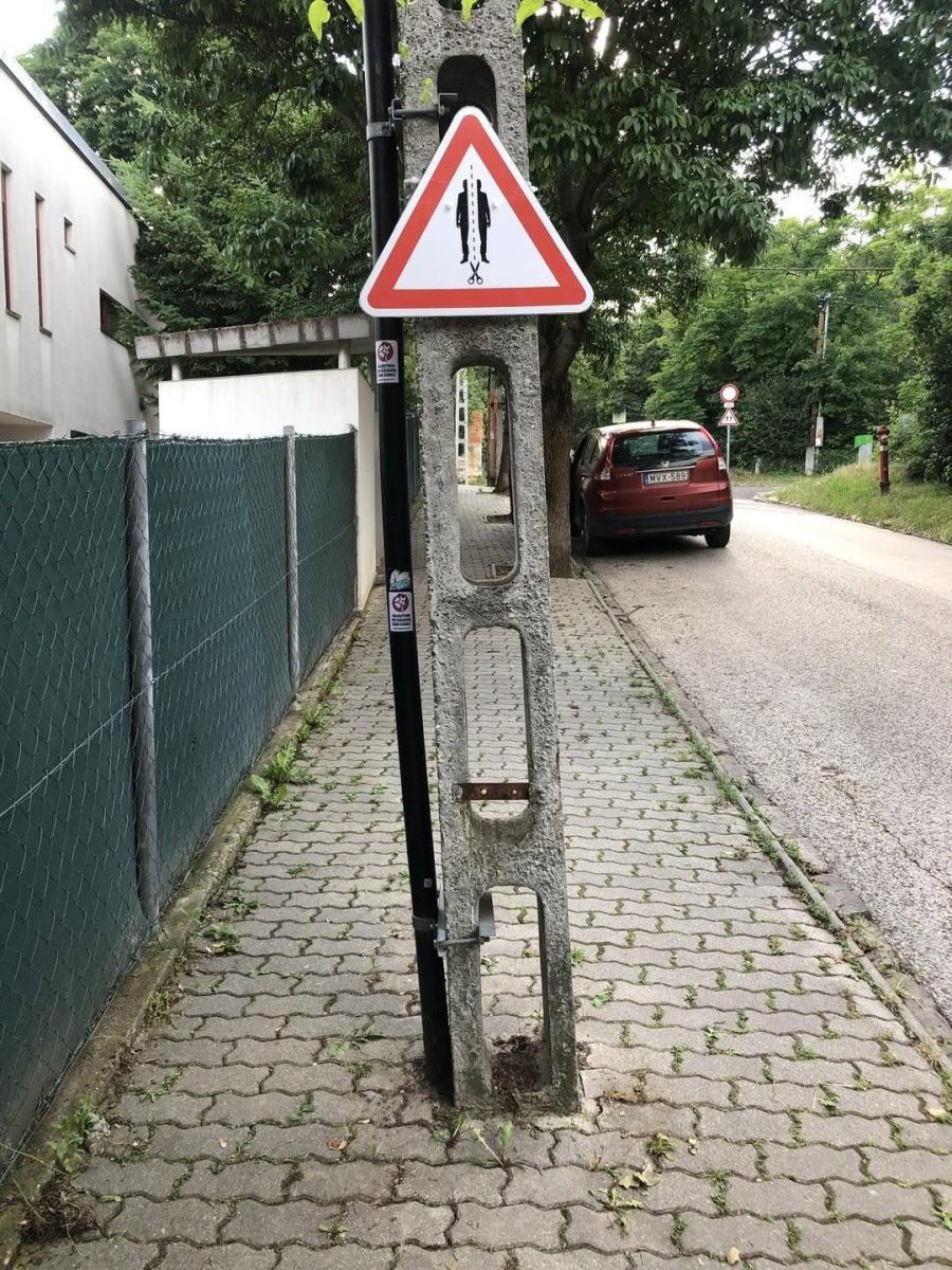 Varovanie pred nebezpečenstvom: lampa s vtipnou značkou na úzkom chodníku vedľa cesty plnej zelene.