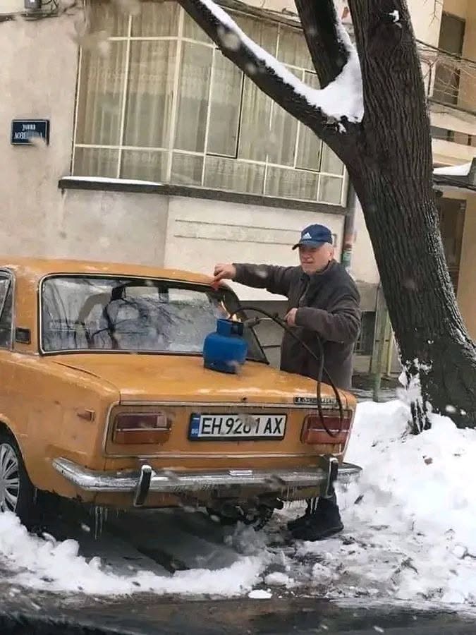 Muž používa horák na rozmrazovanie zadného okna žltého auta v zasneženej ulici.