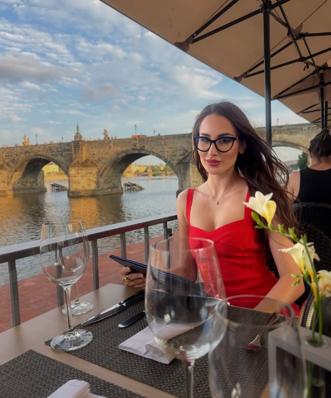 Žena v červených šatách sedí pri rieke s historickým mostom v pozadí, pripravená na večeru na terase.