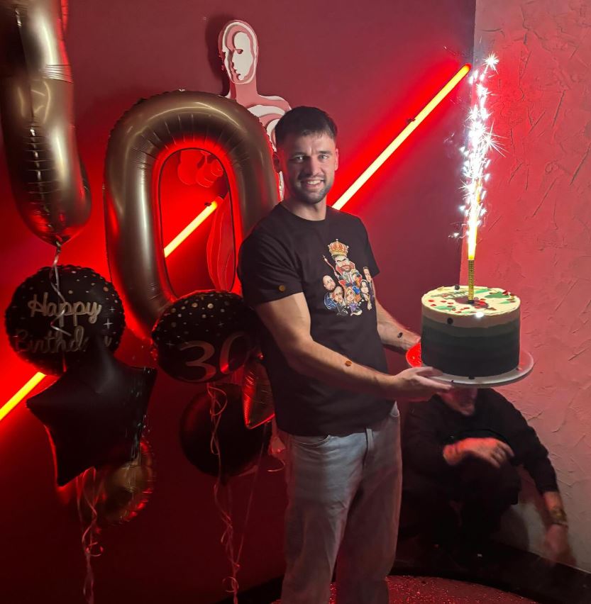 Muž oslavuje 30. narodeniny s tortou a prskavkou, obklopený narodeninovými balónmi v tmavej miestnosti.