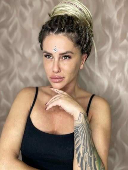 Žena so zložito upravenými vrkočmi a tetovaním na ramene, pózuje s ozdobou na čele pred textúrovaným pozadím.