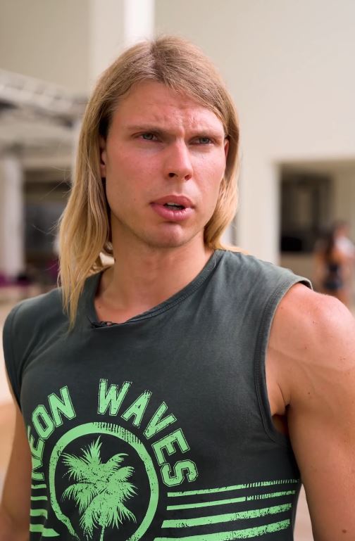 Muž s blond vlasmi v tričku s nápisom Neon Waves stojí vonku pri budove, vyzerá premýšľajúco.
