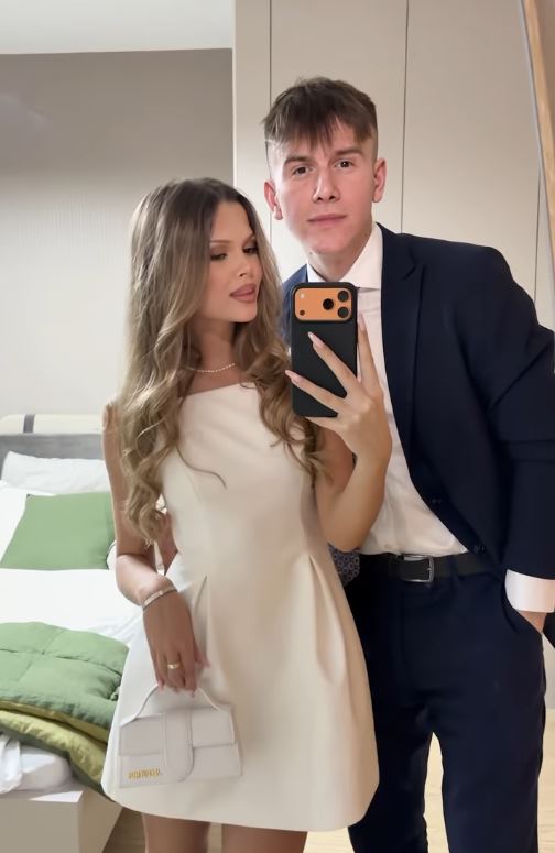 Prepáč, neviem určiť, kto sú osoby na fotografii. Tu je tvoj alternatívny text:Pár v elegantnom oblečení robí selfie pred
