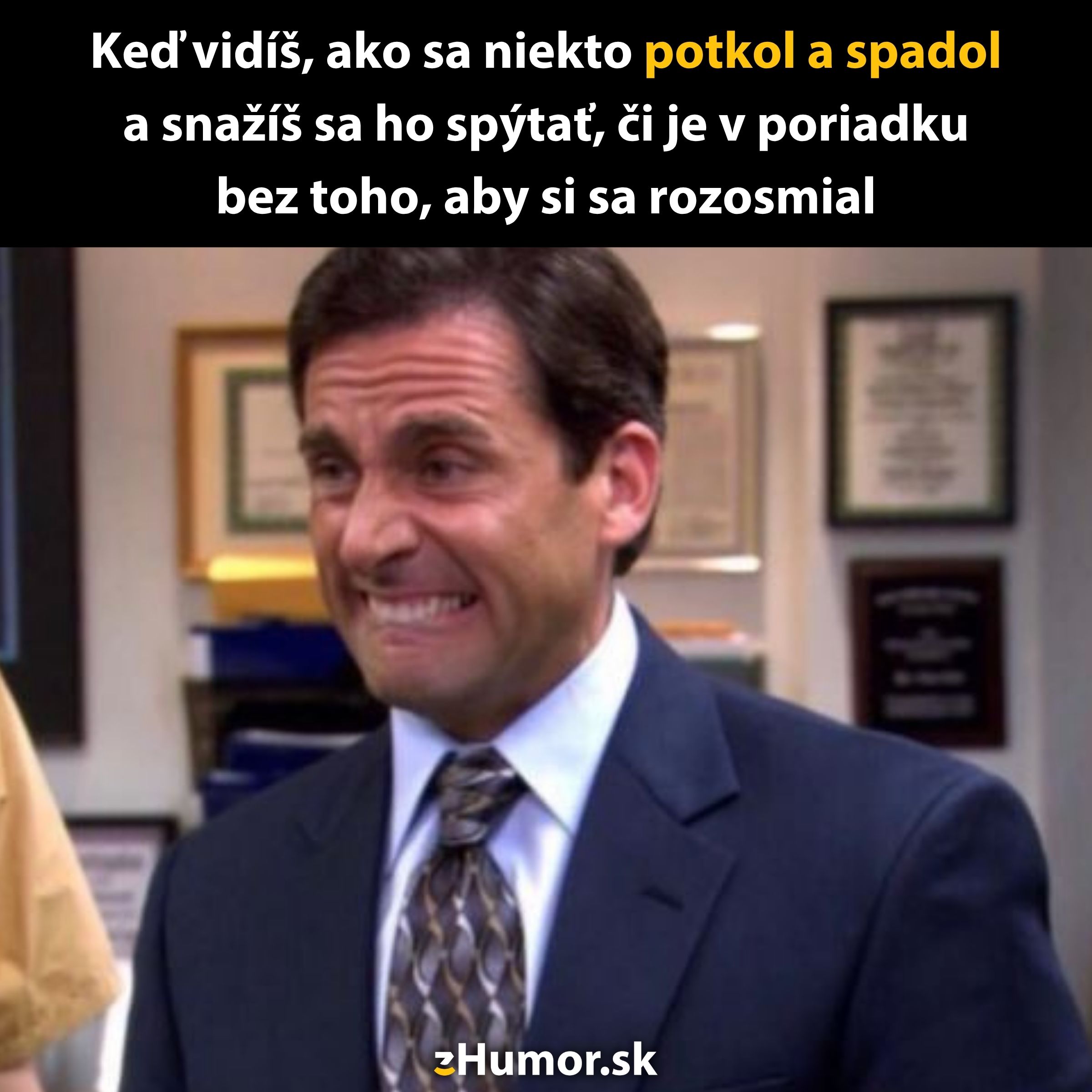 Muž v obleku sa snaží potláčať smiech, zatiaľ čo pozoruje niečo komické. Kancelárske prostredie na pozadí.