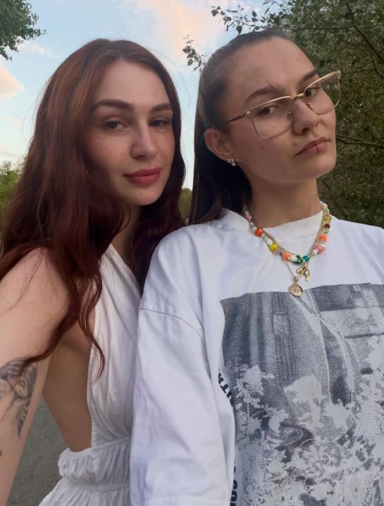 Dve osoby pózujú vonku pri západe slnka, jedna má biely outfit a druhá tričko s potlačou a náhrdelník.