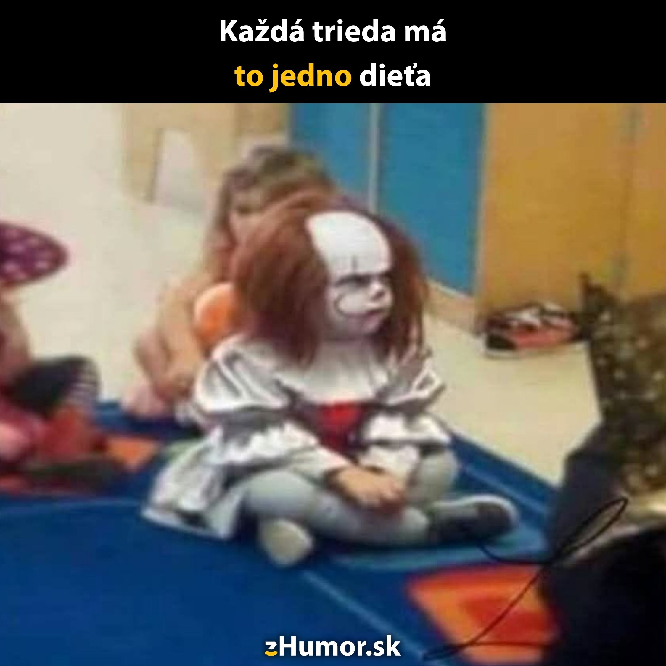 Dieťa v strašidelnom kostýme klauna sedí na koberci v triede, ostatní žiaci ho sledujú. Text: Každá trieda má to jedno dieťa.