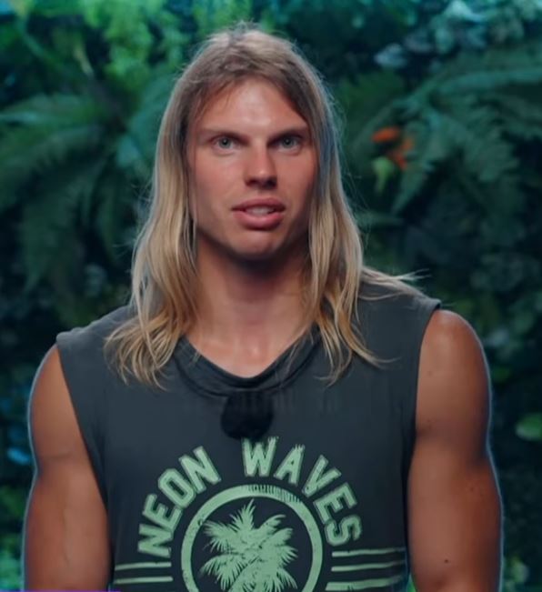 Muž s dlhými vlasmi vo fitnes tílku „Neon Waves“ stojí pred zeleným rastlinným pozadím.