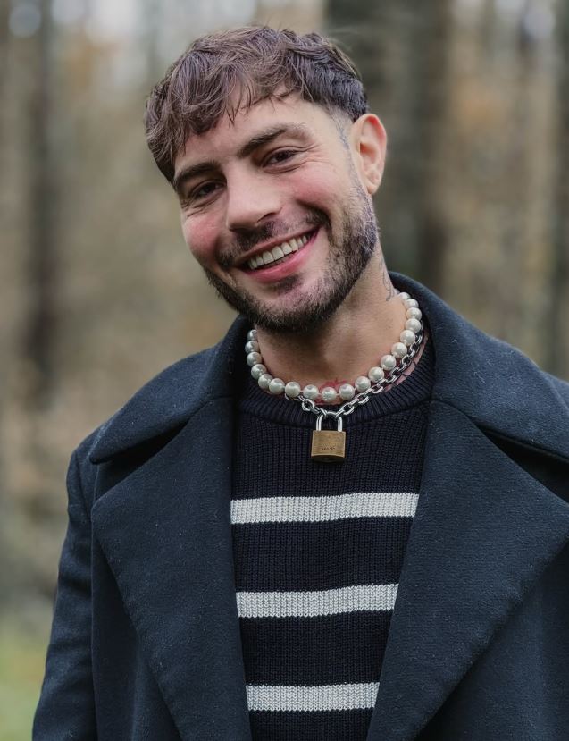 Usmievavý muž v tmavom kabáte s výrazným náhrdelníkom, stojaci v prírodnom prostredí.