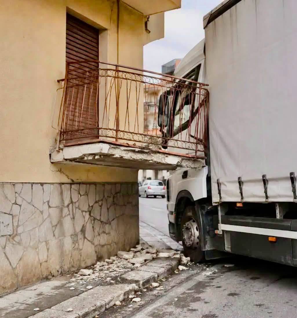 Nákladné auto naráža do balkóna na úzkej ulici; poškodená fasáda budovy a trosky na chodníku.